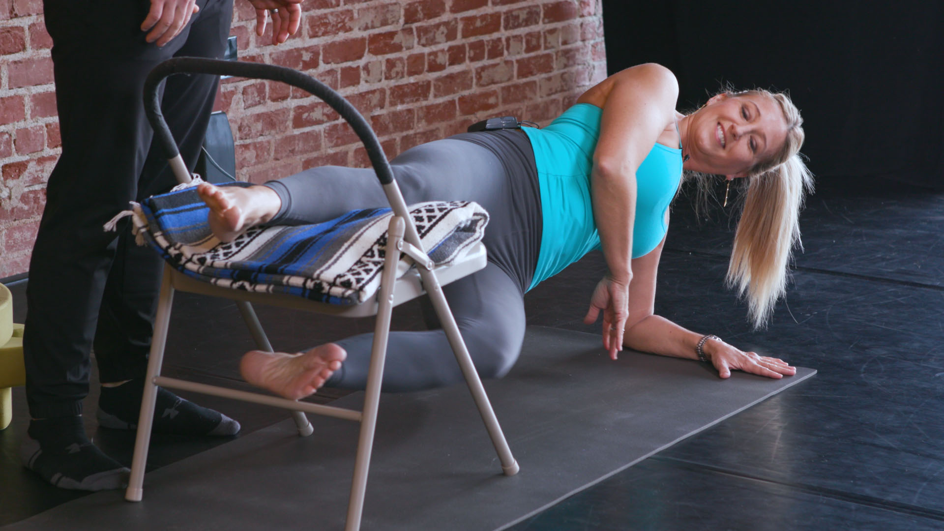 A+ Adductor Jules on her side with the top leg on a chair, pressing down to hover the bottom leg. The rest of her body is off the floor with the exception of the bottom forearm.