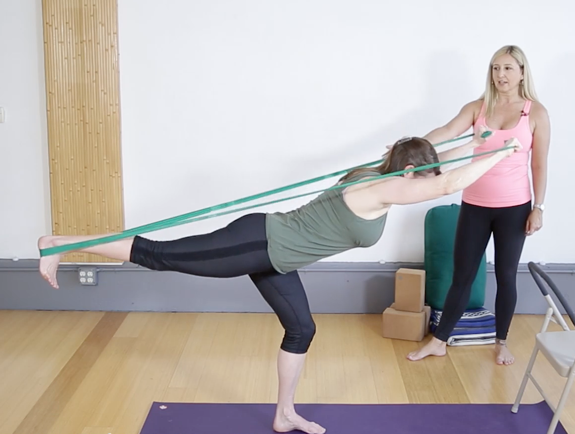 Jules instruction Catherine in Warrior 3 pose with a theraband around her lift foot and pulling it taught with the arms overhead.