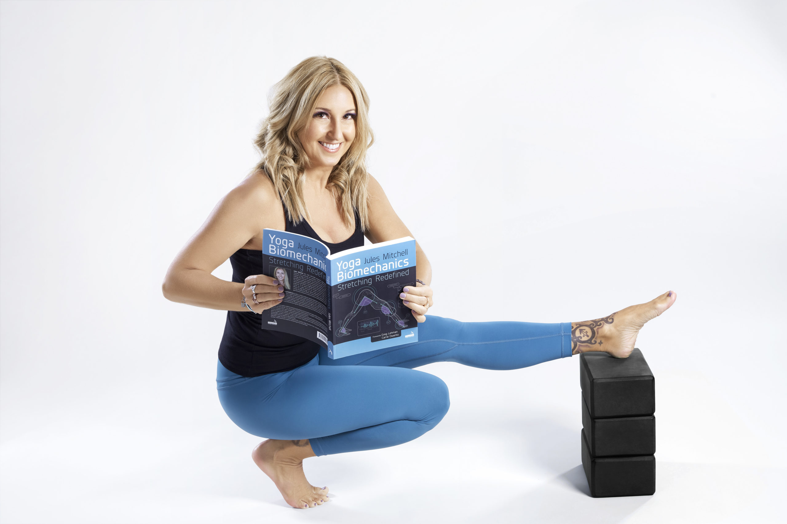 Jules is holding her book and smiling with one leg resting on some yoga blocks.